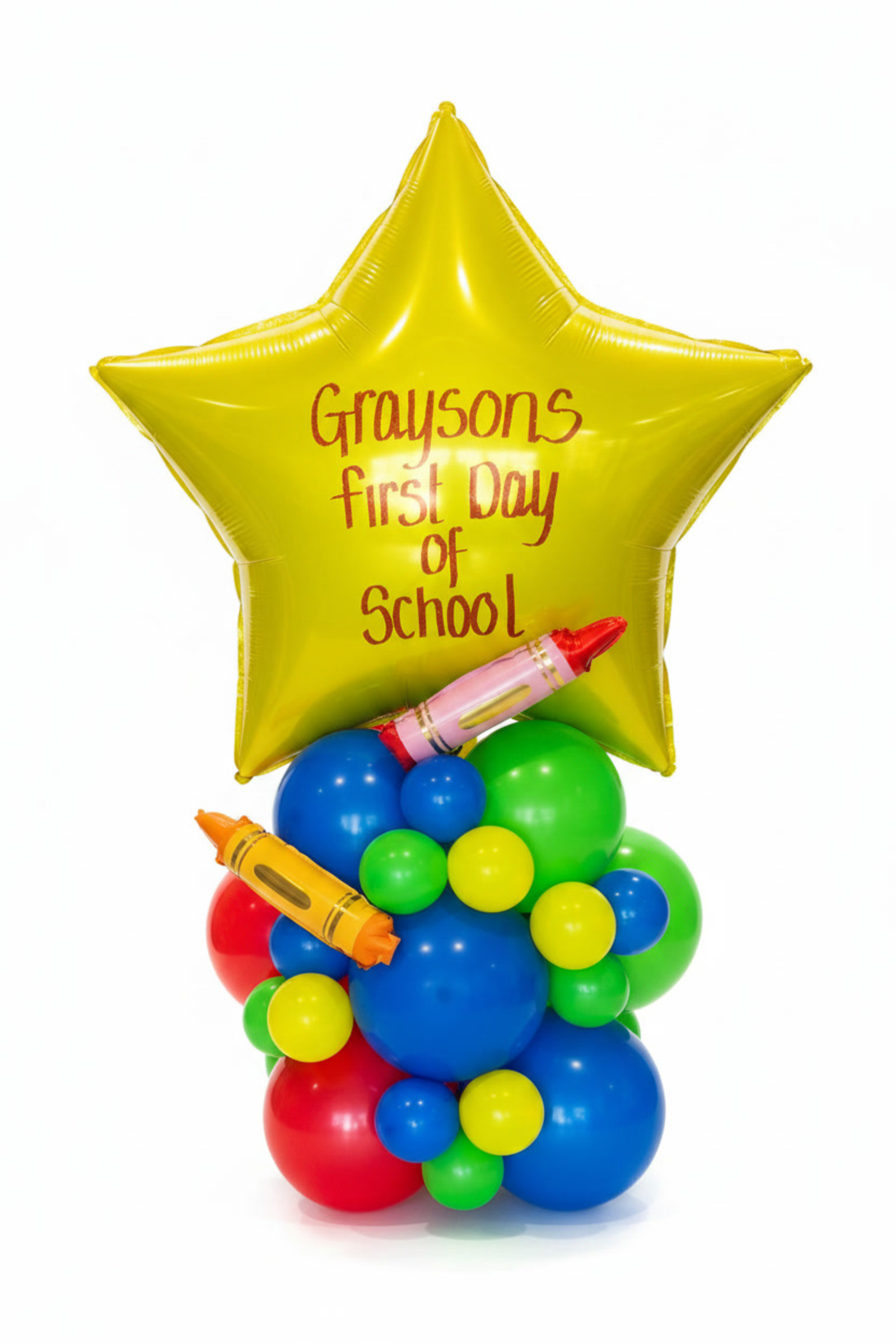 A first day of school balloon bouquet featuring a large star balloon with the text 'Grayson's first day of school' in orange, mounted on a cluster of red, blue, green, and yellow balloons, with crayons resting on top, set against a white background.