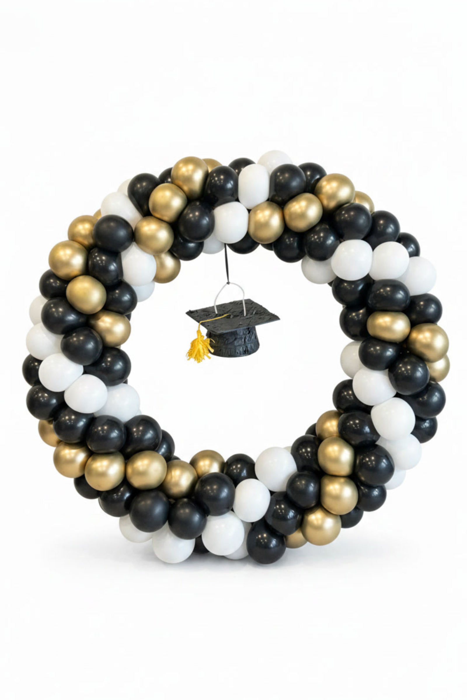 A graduation-themed balloon wreath made of black, white, and metallic gold balloons, with a black graduation cap and tassel hanging in the center, on a white background.